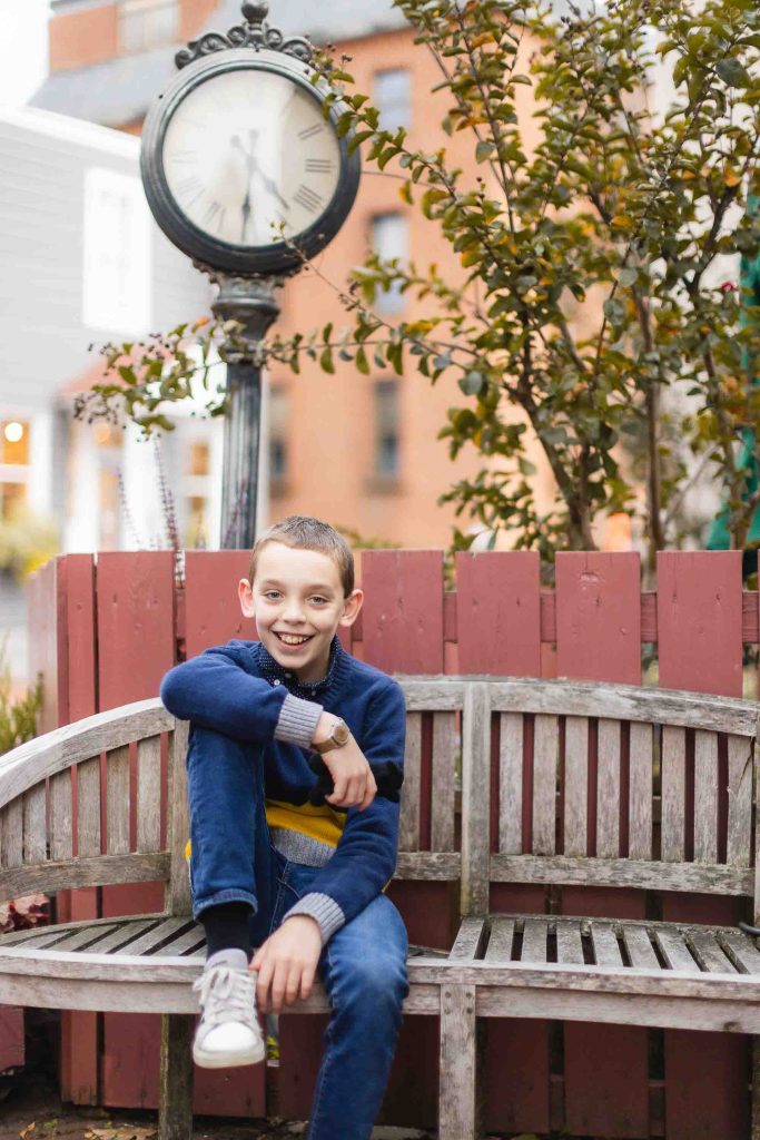 A boy sits on a wooden bench in front of a clock in Downtown Annapolis.
