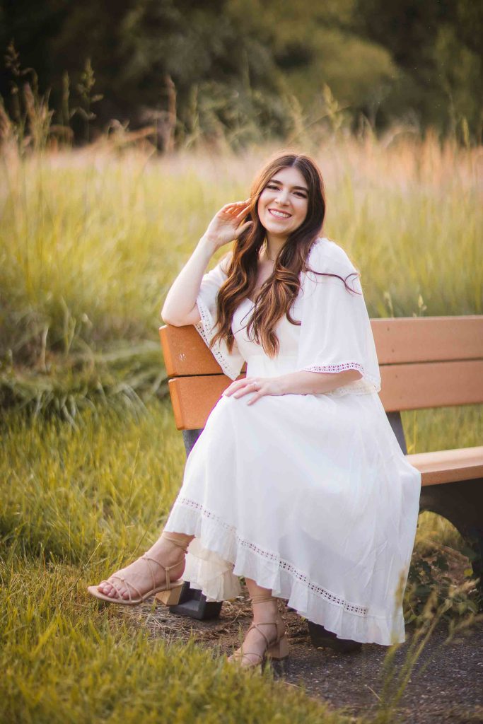 A woman in a white dress sitting on a bench in the beautiful Patuxent Research Refuge field, Maryland, indelibly capturing the essence of timeless elegance for her engagement photos.