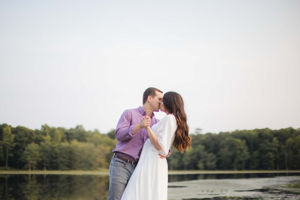 An engaged couple sharing a romantic kiss during a stunning sunset in Maryland, capturing the essence of their engagement photos at a picturesque lake.