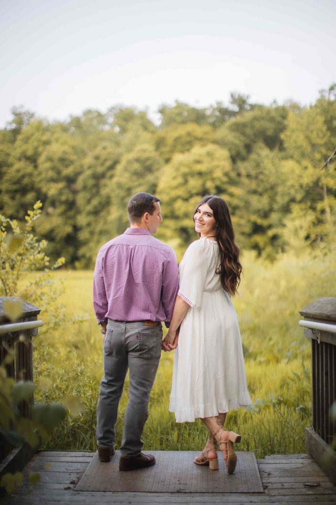 An engaged couple standing on a bridge at Patuxent Research Refuge in Maryland, admiring the beautiful sunset over the woods.