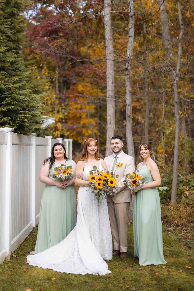 A bride and her bridesmaids pose with sunflowers in front of a white fence at their wedding portrait session.