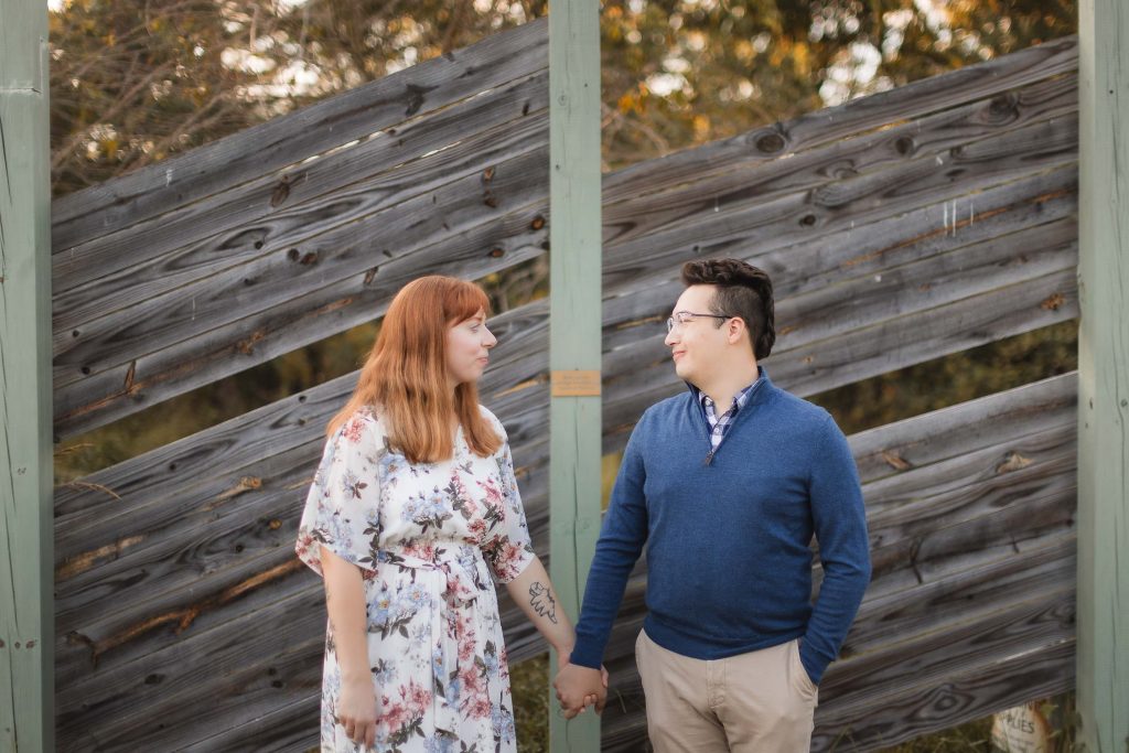 An engagement portrait of a couple holding hands in front of a wooden fence at Patuxent Research Refuge.