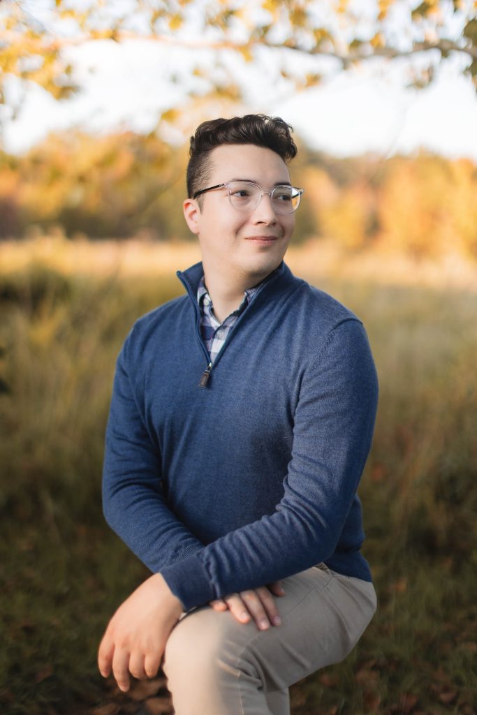 A young man in glasses sitting in a field for an engagement portrait at Patuxent Research Refuge.