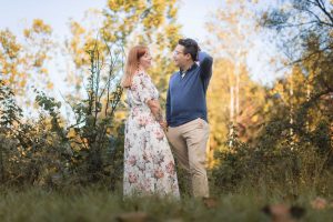 An engaged couple is standing in the middle of a field with trees in the background, capturing stunning engagement portraits at Patuxent Research Refuge.