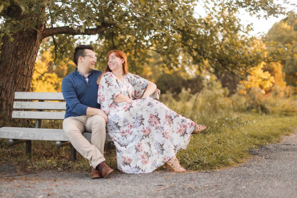 An engagement couple sits on a bench under a tree at Patuxent Research Refuge while taking portraits.