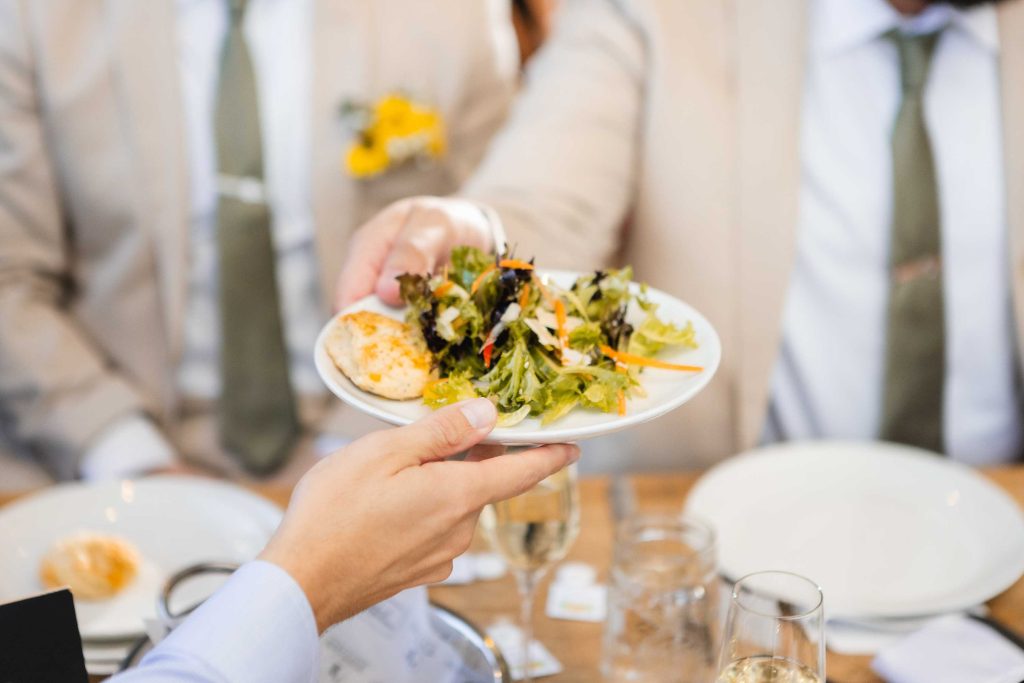 At a wedding, a man is handing a plate of salad to another man with intricate details.