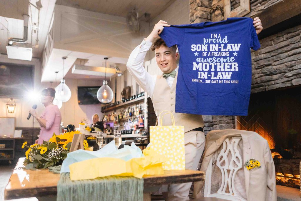 A man is holding up a t-shirt that says "I'm a sorority sister and a mother" at a wedding reception at Blackwall Barn & Lodge.