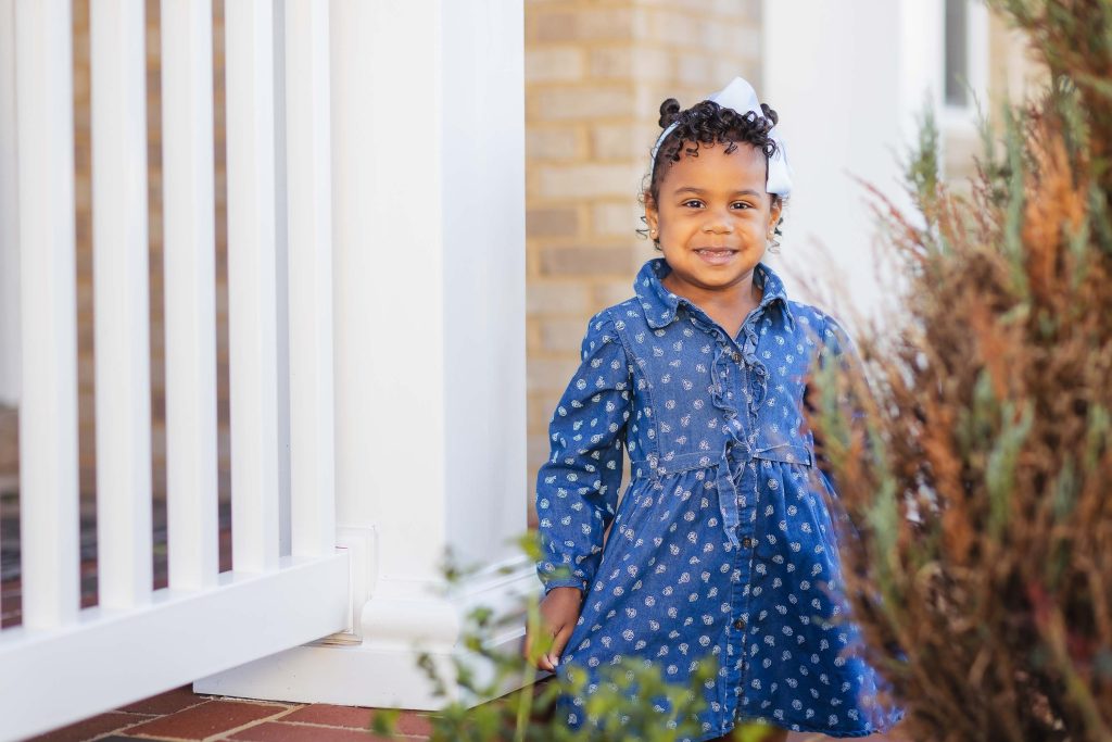 A little girl in a blue dress standing in front of a white fence, captured in a family portrait at home.