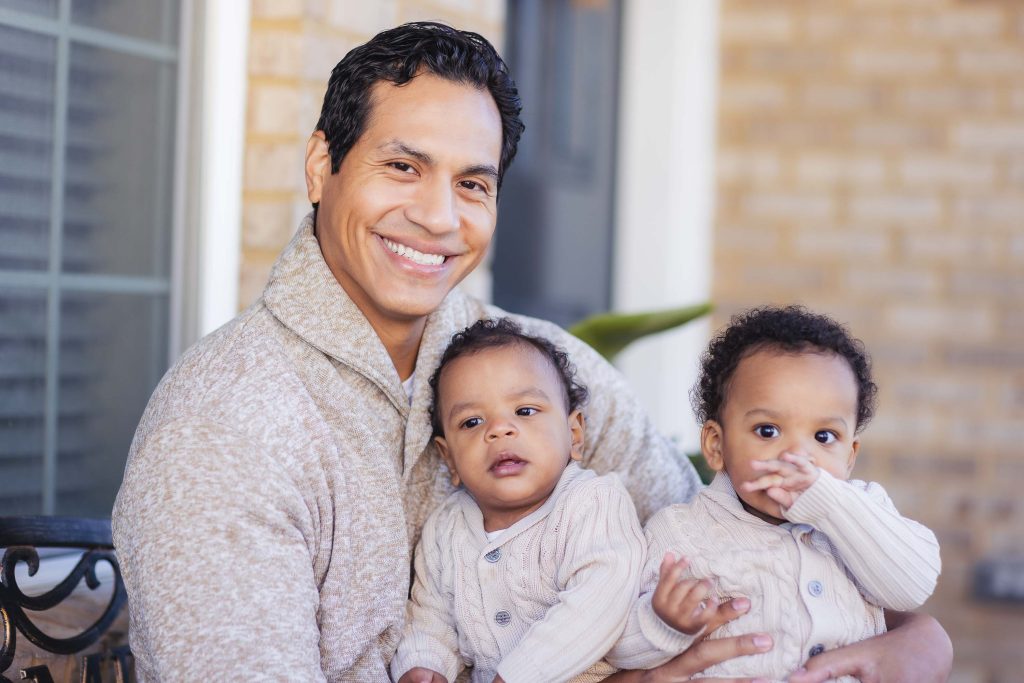 A man holding his two young children for a family portrait at home.