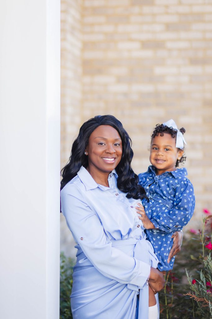A pregnant woman at home holding her daughter in front of a brick wall, capturing a heartfelt family portrait.