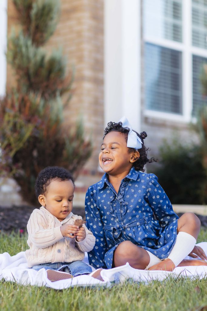 Two black children at home sitting on a blanket in front of their house.