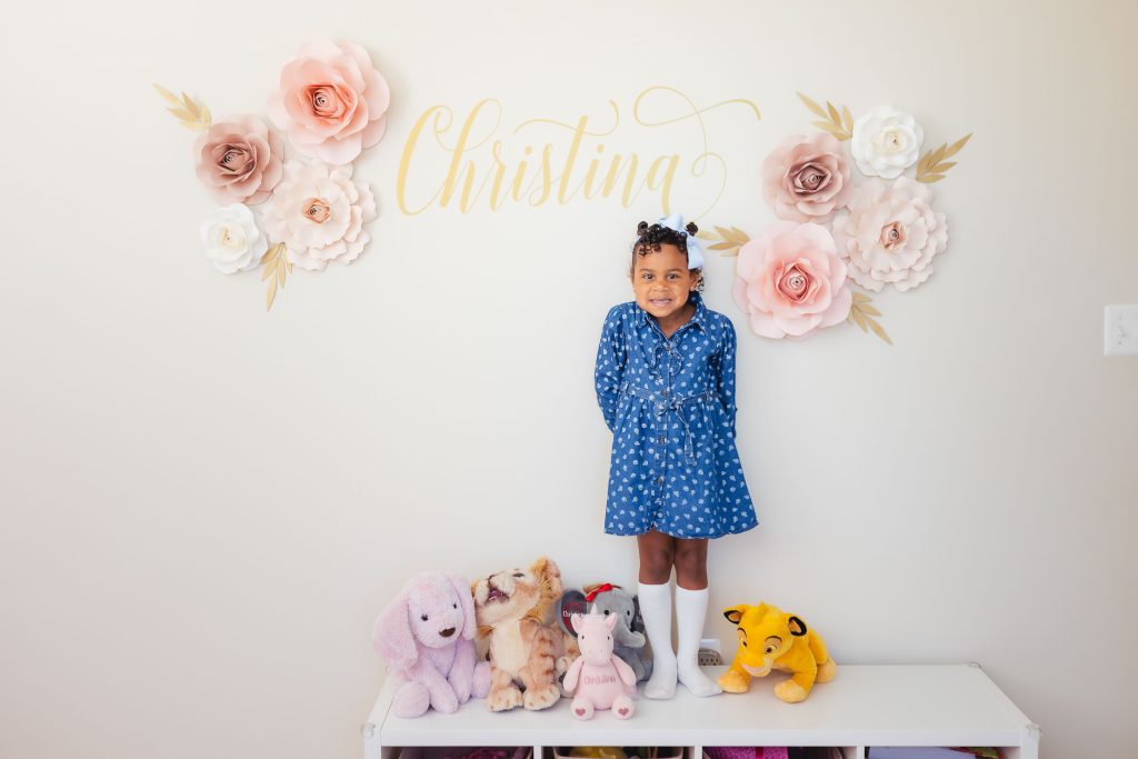 A little girl standing at home in front of a wall adorned with flowers and surrounded by stuffed animals.