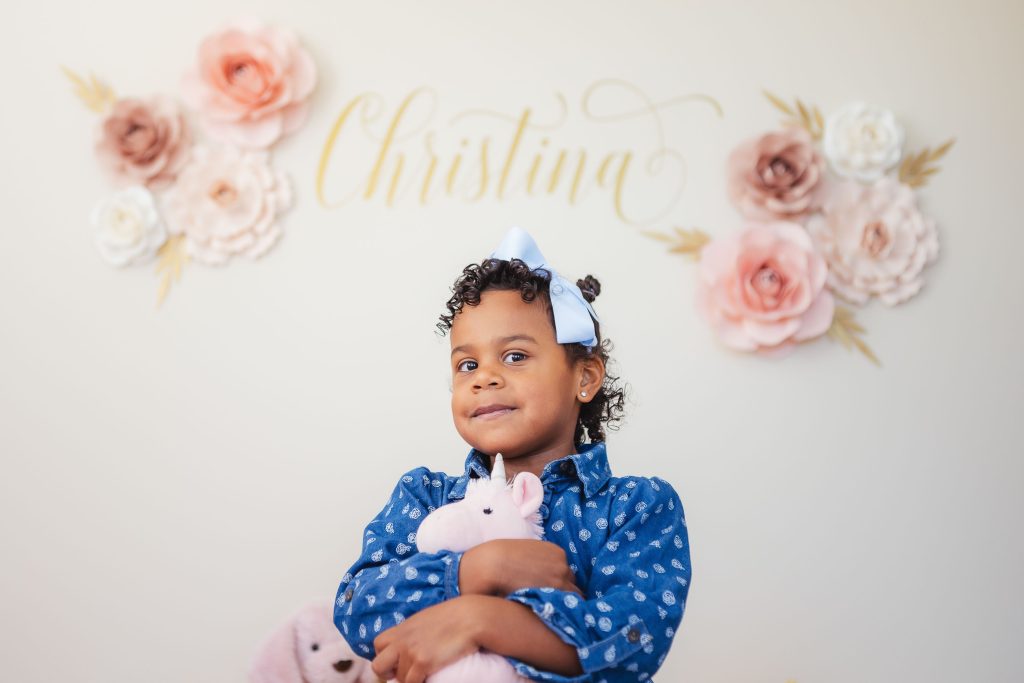 A little girl holding a pink teddy bear in front of a flower wall. Home Portraits.