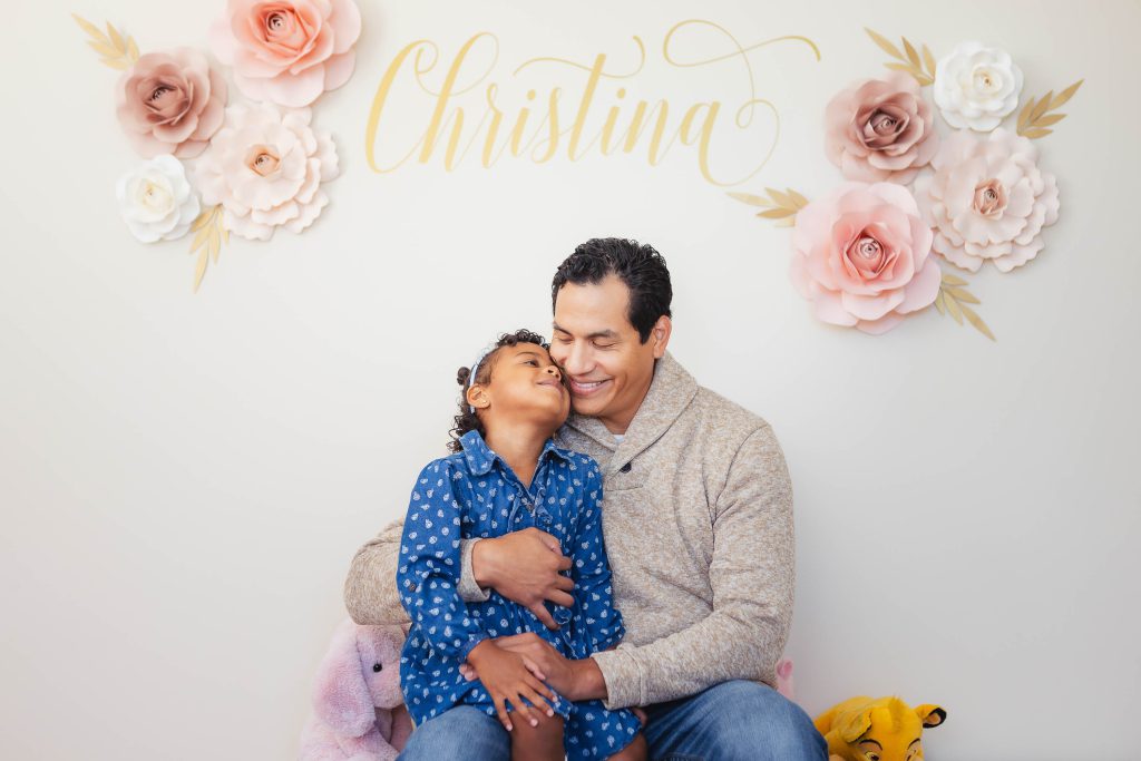 A family portrait captured at home, featuring a man and a little girl sitting in front of a stunning flower wall.