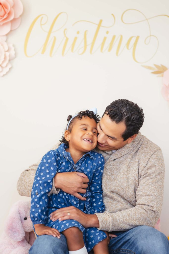 A man and a little girl hugging in front of a flower wall in a heartwarming family portrait.