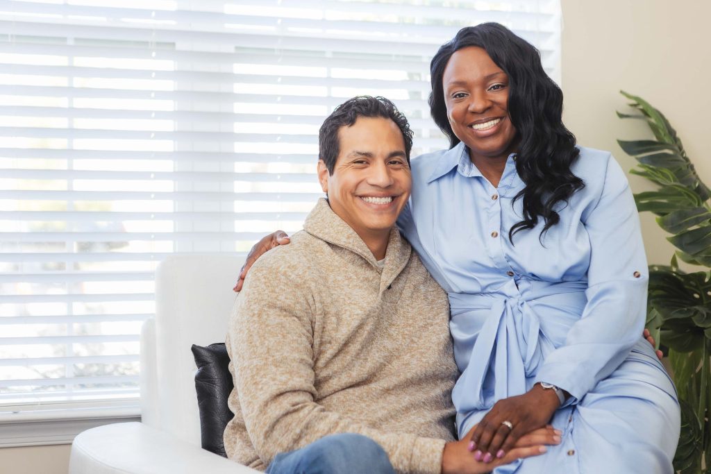 A black man and woman sitting on a white couch, capturing intimate family portraits at home.