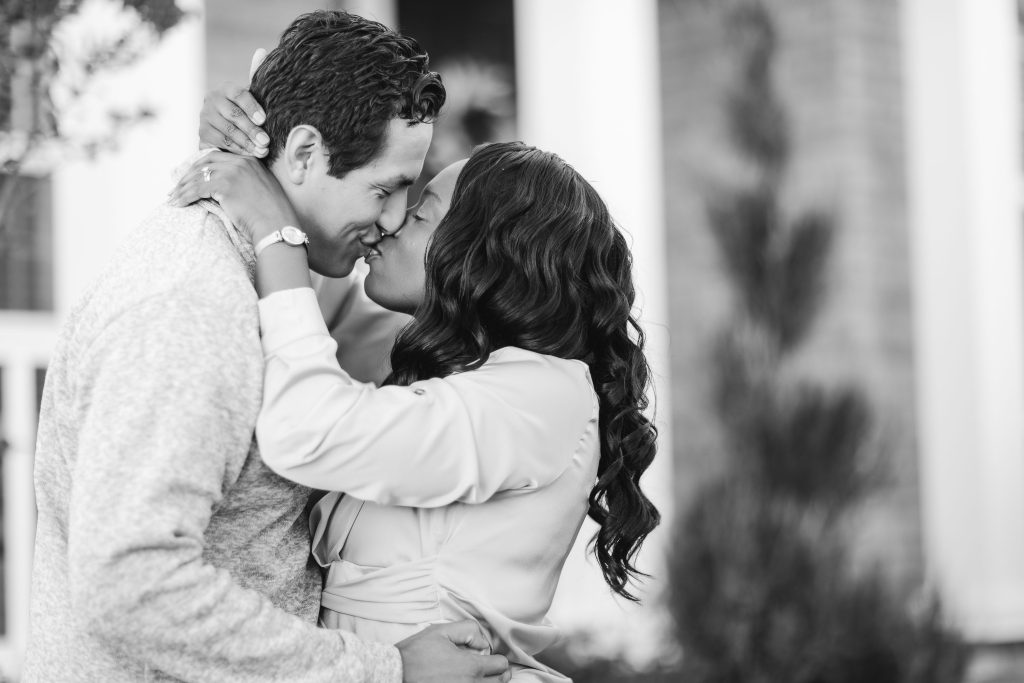 A black and white photo of a couple kissing at home in front of a house.