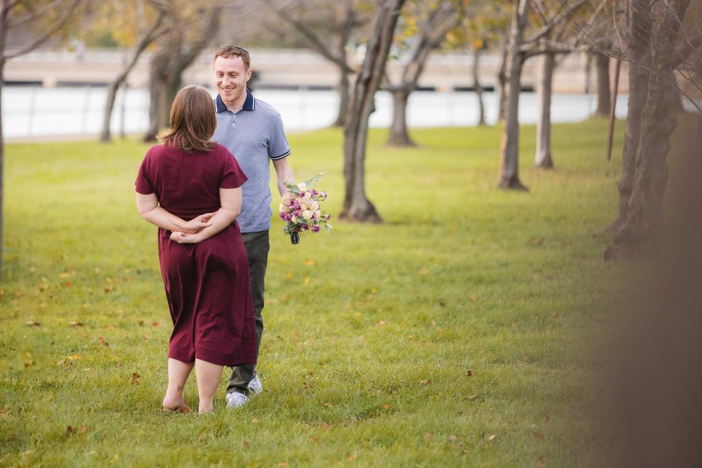 A man and woman are walking in the grass near a lake, filled with surprise as he plans a wedding proposal.