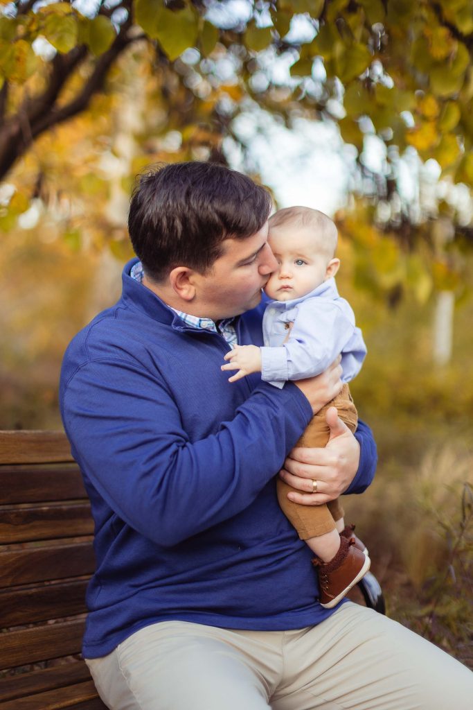 A man kisses his baby on a bench at Quiet Waters Park in the fall.