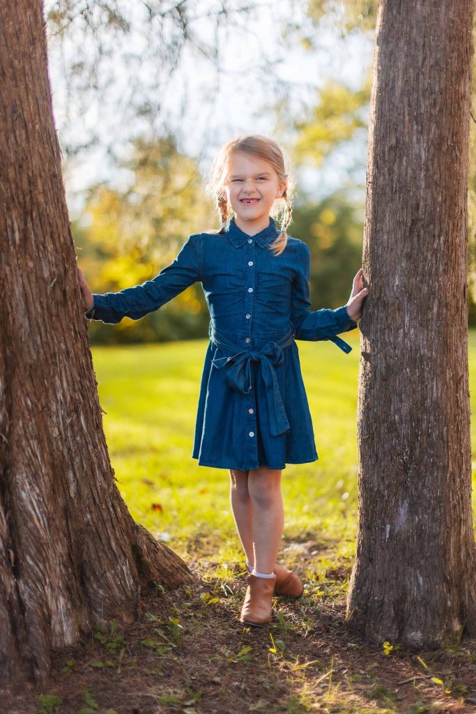 A little girl in a blue dress posing for portraits against a tree in Bowie, Maryland.