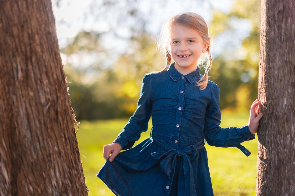 A little girl in a denim dress posing for a family portrait against a tree at Belair Mansion.