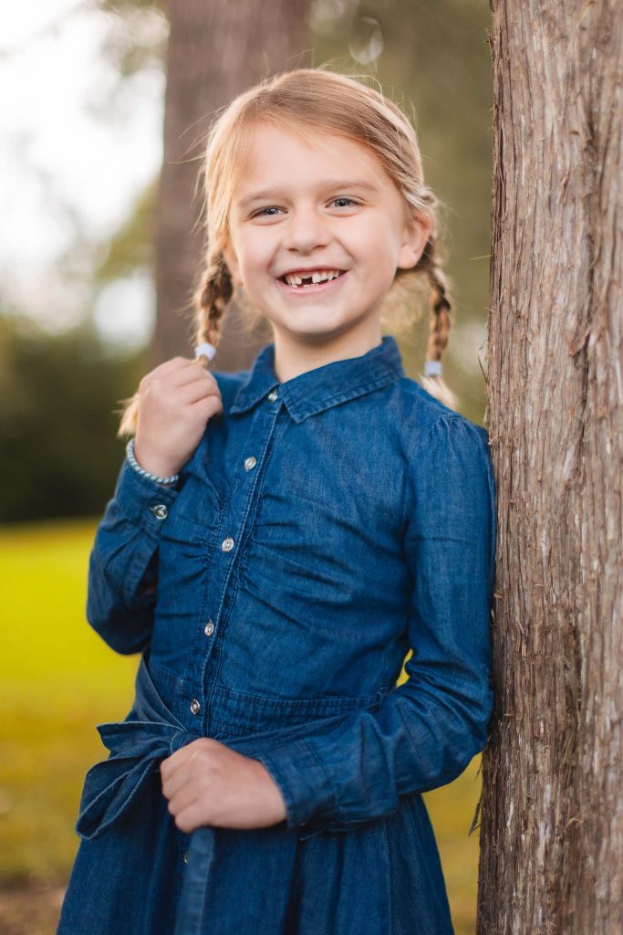A little girl in a denim dress leaning against a tree in Bowie Maryland for family portraits.