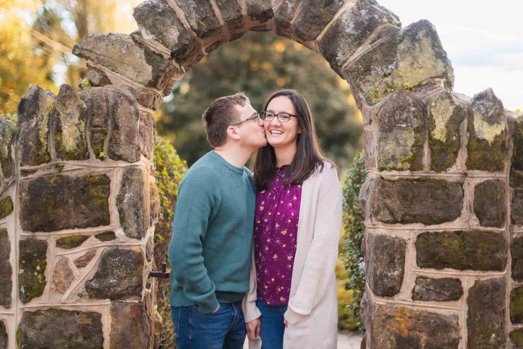 A couple kisses during their engagement session in front of a stone archway in Bowie, Maryland.