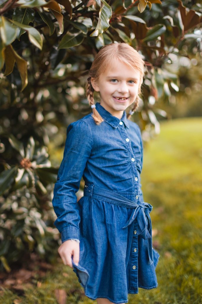 A little girl wearing a denim dress poses for family portraits in front of a tree in Bowie, Maryland.