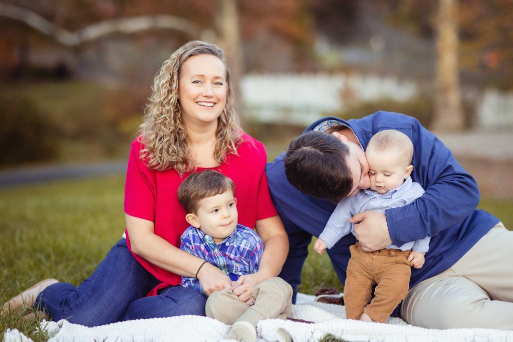 A family sits on a blanket in Quiet Waters Park, Annapolis Maryland and kisses their children during a portrait session.