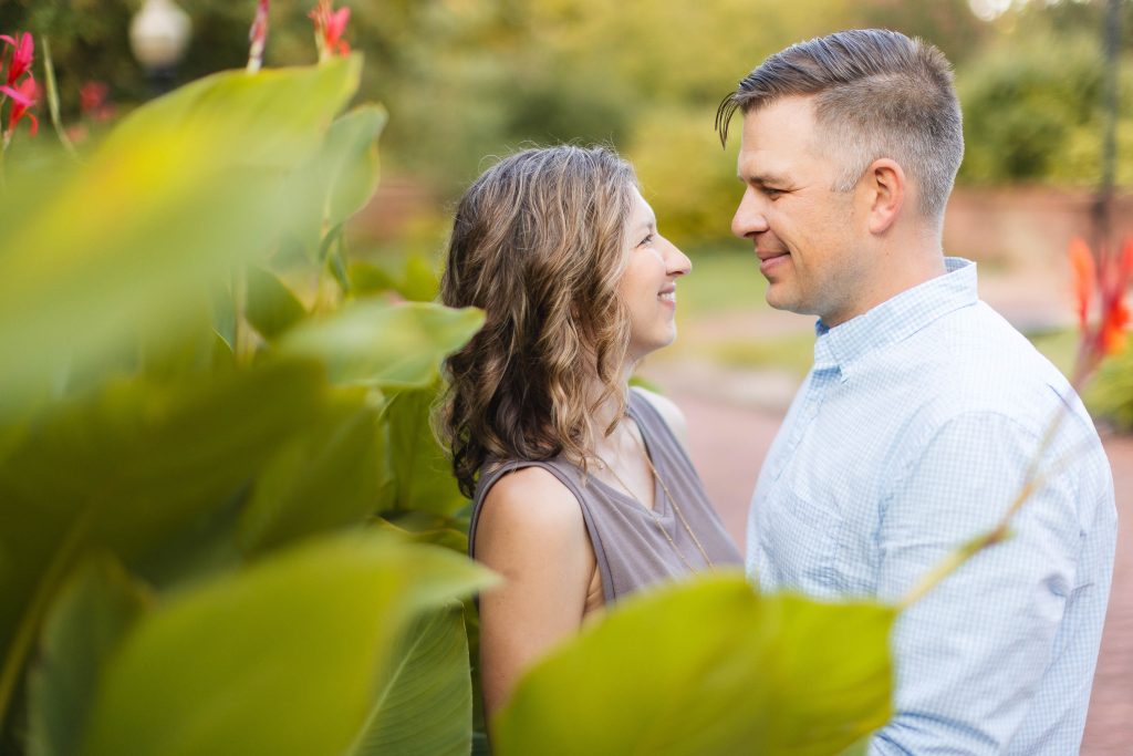 A smiling engaged couple poses for their portrait at Quiet Waters Park in front of lush bushes.