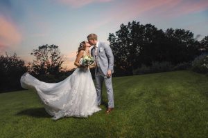 A bride and groom kissing in the grass at sunset.