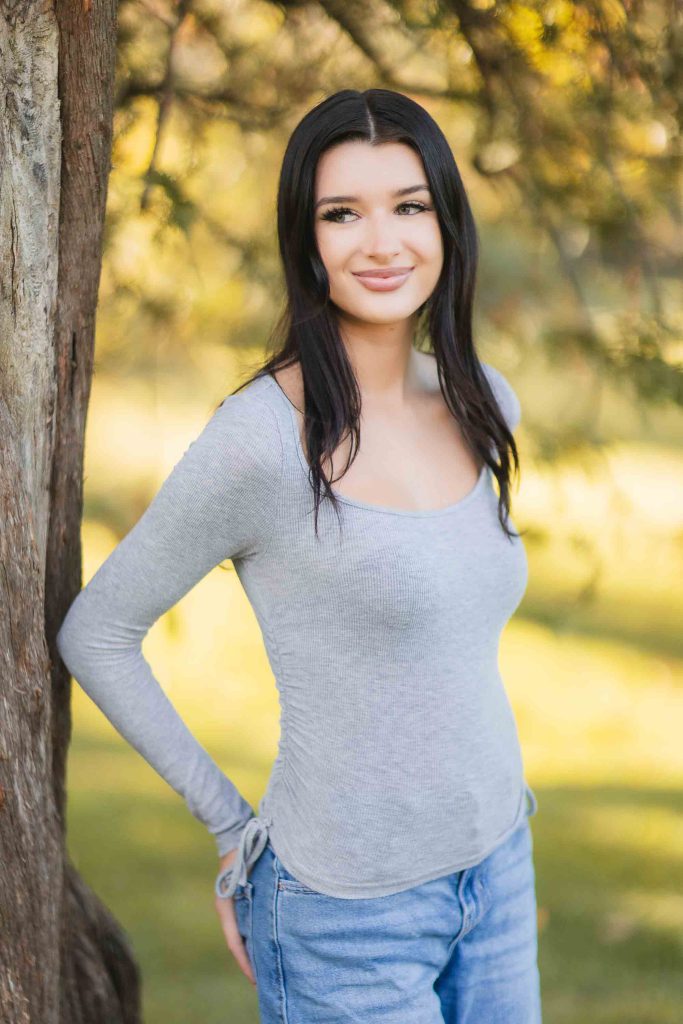 A woman with long dark hair is standing outdoors beside a tree in Bowie, Maryland, wearing a light grey long-sleeve shirt and blue jeans. The background features greenery and sunlight, capturing the essence of the mini-session perfectly.