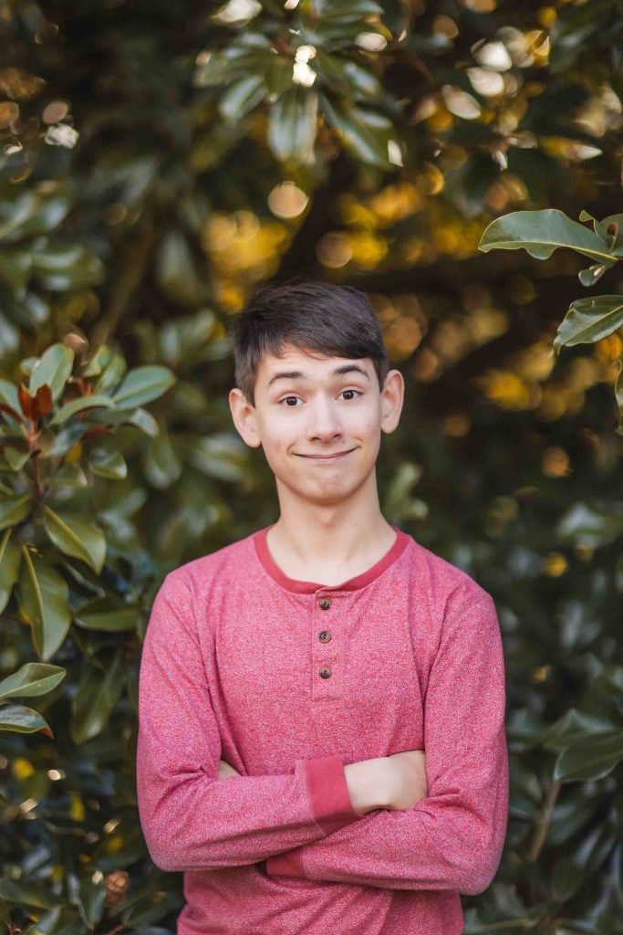 A young person with short dark hair, wearing a red long-sleeve shirt, stands outdoors at Belair Mansion in Maryland with arms crossed and a playful facial expression. Green leaves create a lush background for this charming portrait.