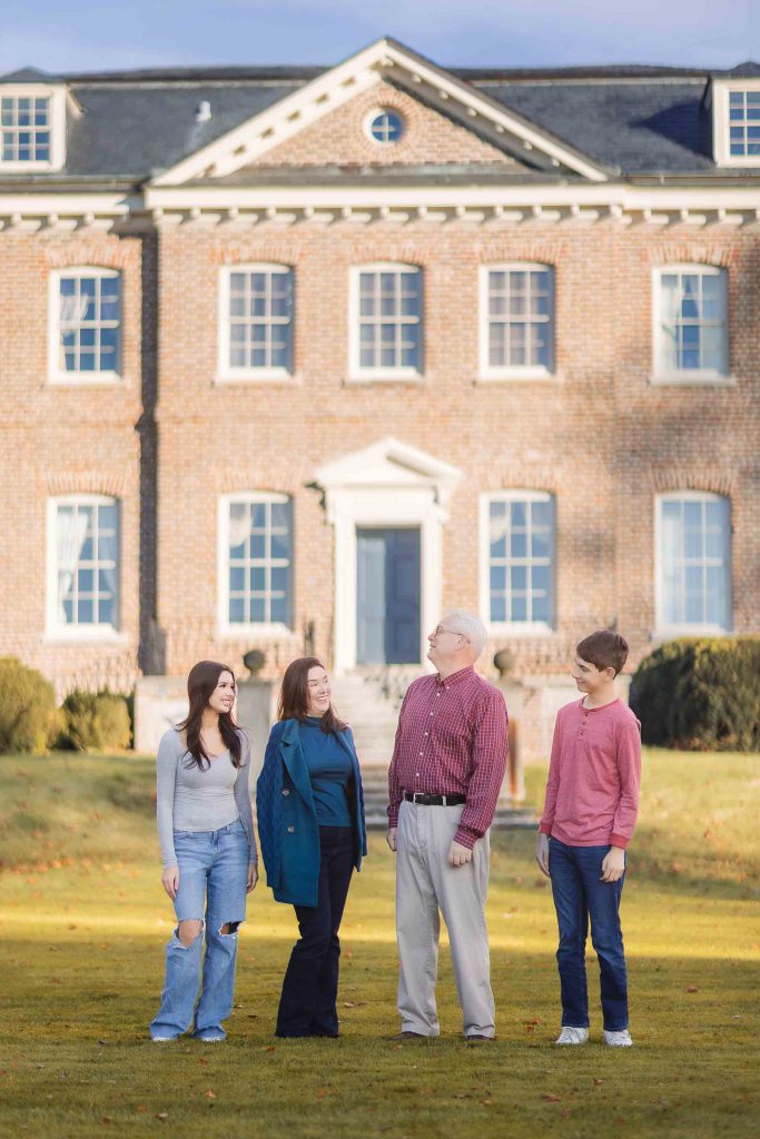Four people stand in front of a large, two-story brick building with multiple windows and a central white door, capturing the moment as if it were a Bowie-inspired portrait from a mini-session.