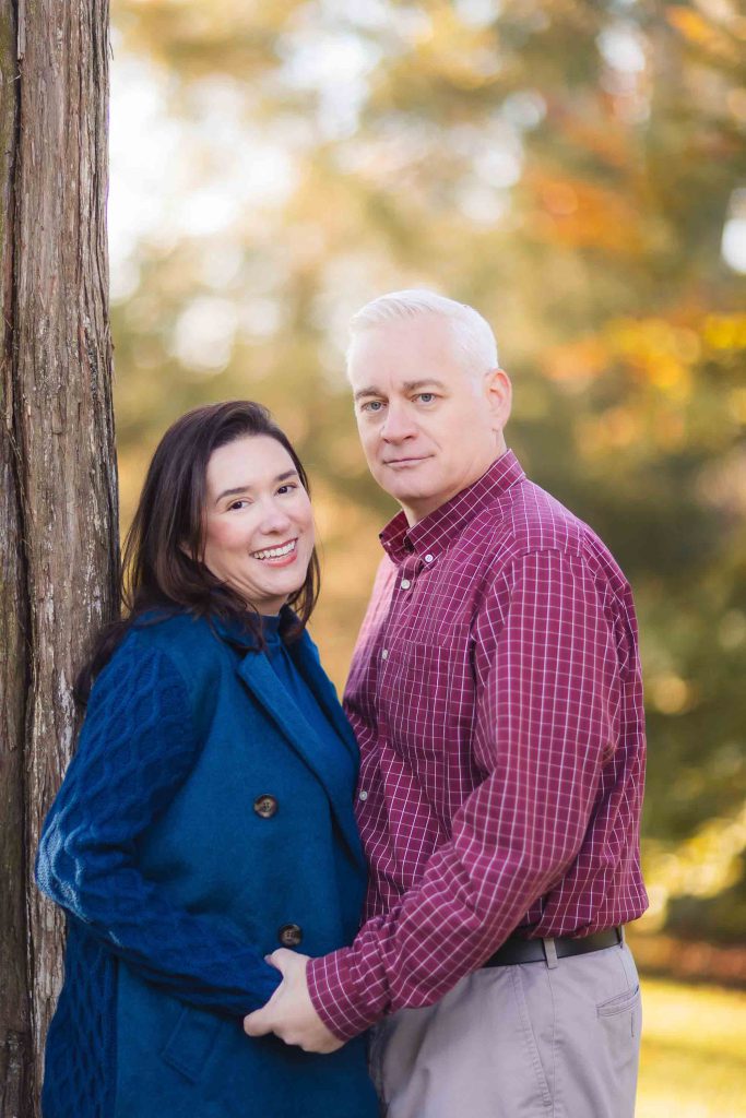 A man and woman stand outdoors holding hands, the woman smiles at the camera while the man looks ahead. They are dressed in casual autumn attire, perfect for a Bowie family mini-session. Trees with fall foliage are in the background.