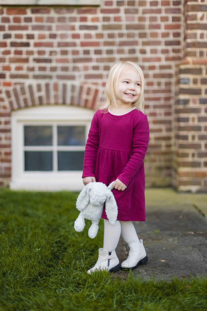 A young girl in a maroon dress stands outside in front of a brick building, holding a plush white rabbit. She is smiling and looking to her left, capturing a perfect moment for this family portrait mini-session.