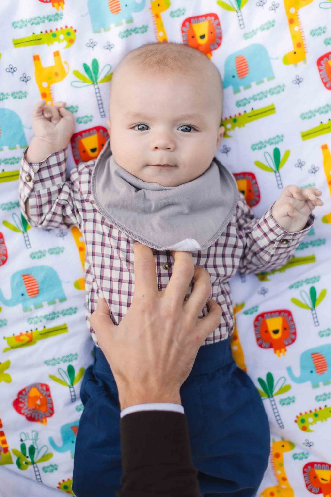 During a charming mini-session in Maryland, a baby wearing a checkered shirt and a bib lies on a colorful blanket with animal prints as an adult hand gently touches the baby's chest, capturing the perfect portrait moment.