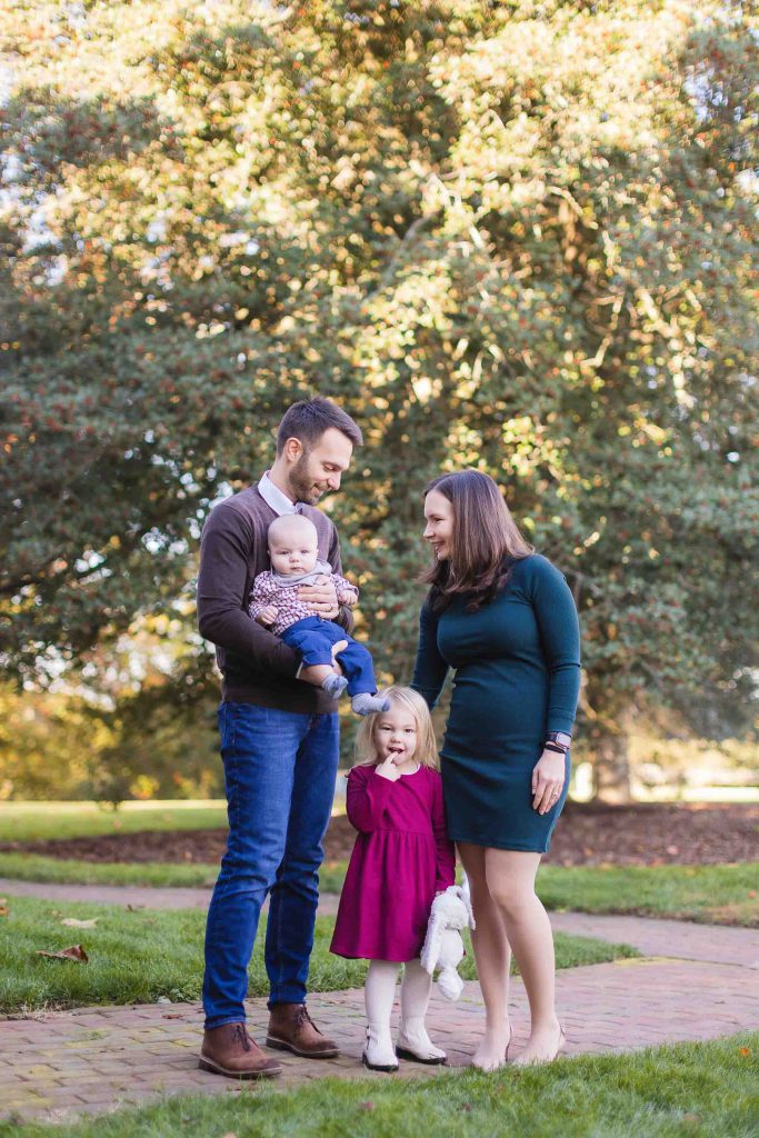 A man and woman stand on a walkway in a park in Bowie, Maryland, with the man holding a baby and the woman next to a young girl in a red dress. Trees and greenery provide a beautiful backdrop for this delightful mini-session.