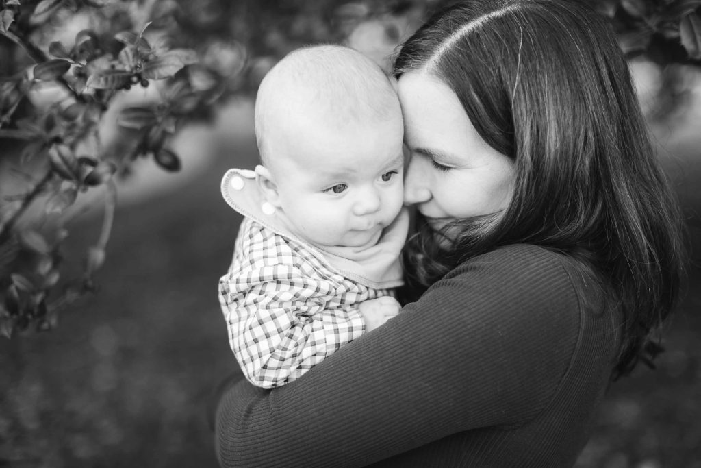 A woman holds a baby close, with the baby's face resting against her cheek, in a tender embrace. The black and white portrait captures this intimate moment against a blurred background of leafy branches. This heartwarming mini-session took place in Maryland.