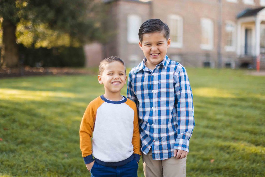Two young boys stand close together and smile in this charming portrait taken outdoors on a grassy area, with a building and trees in the background during their mini-session.