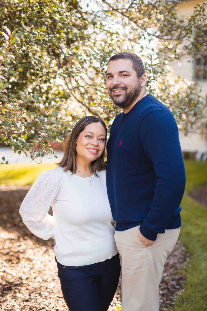 A man and woman stand closely together outdoors near a bush at Belair Mansion, smiling at the camera. The man wears a navy sweater and beige pants, while the woman wears a white sweater and dark pants. This lovely moment was captured during their mini-session.