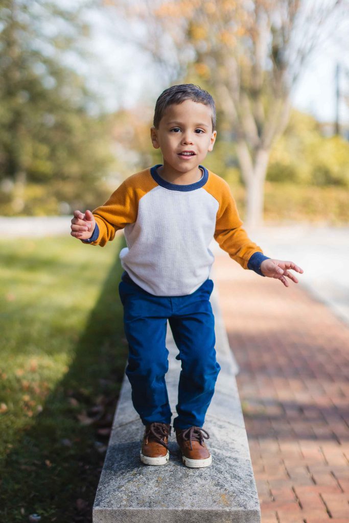 A young boy in a white and orange long-sleeve shirt and blue pants stands on a low concrete ledge outdoors, appearing to balance himself with extended arms, reminiscent of carefree days spent exploring the grounds of Belair Mansion.
