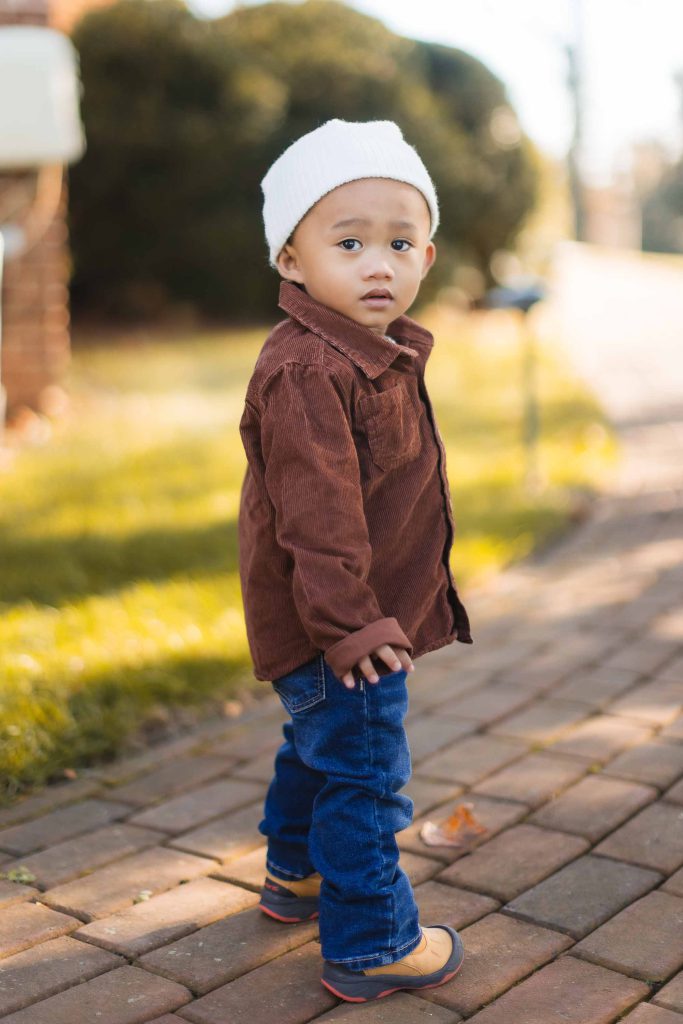 A young child wearing a white beanie, brown jacket, blue jeans, and brown shoes stands on a brick path at Belair Mansion, looking over their shoulder. The mini-session captures the essence of curiosity with a grassy area and blurred background visible.