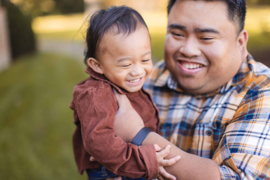 A joyful adult and a child smile, with the child held in the adult's arms. The adult wears a plaid shirt and the child wears a brown outfit. They are outdoors at Belair Mansion, Maryland, with blurred greenery in the background during their mini-session.
