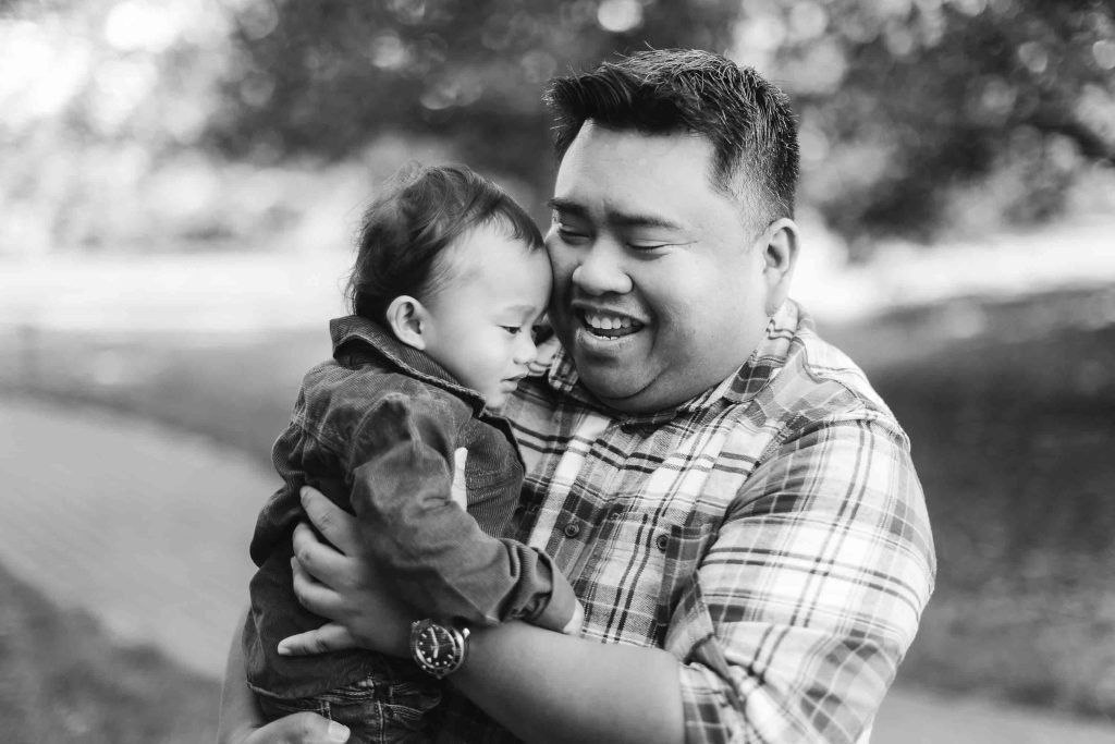A smiling man in a plaid shirt holds a baby in his arms outdoors during a portrait mini-session. Both appear to be content and happy. The background shows blurred trees and a pathway, capturing the serene beauty of Maryland.