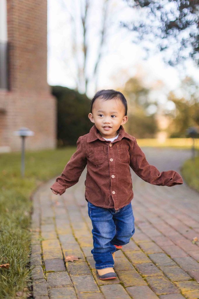 A young child with short dark hair walks down a brick path in Bowie, Maryland. The child is wearing a brown shirt and blue jeans. Trees and a building are visible in the background, showcasing the charm of this family-friendly community.