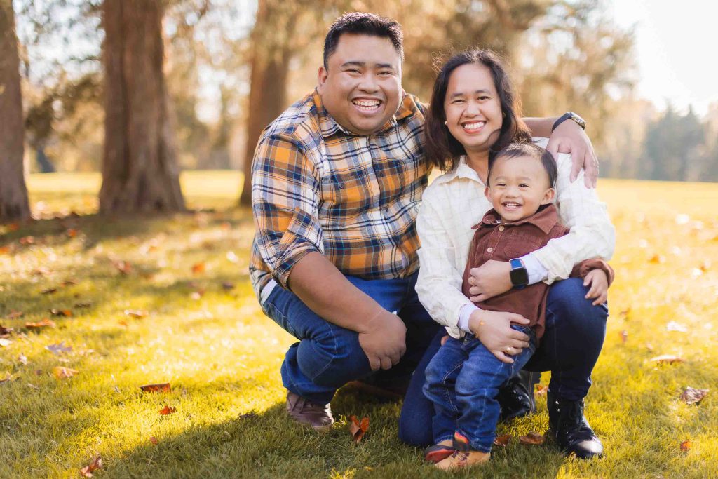 A smiling family of three sits and kneels together on grass in a park, with trees and sunlight in the background. This charming portrait captures a man in a plaid shirt, a woman in a cream shirt, and a child in brown and blue jackets during their mini-session.