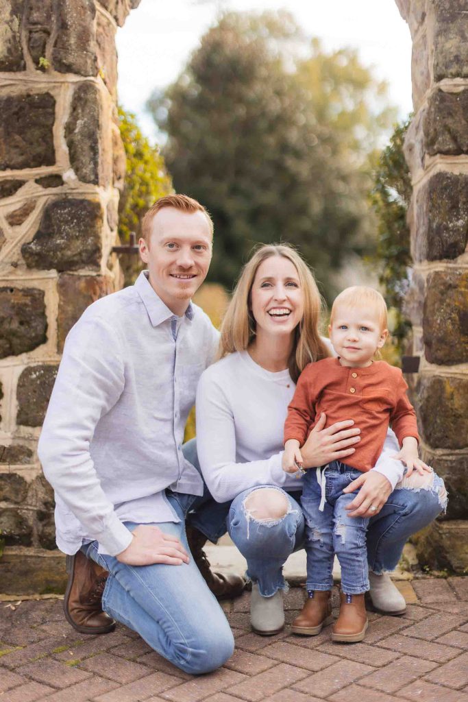 A family of three poses for a mini-session photo outdoors in front of a stone archway in Maryland. The man, woman, and young child all smile warmly, dressed in casual clothing.
