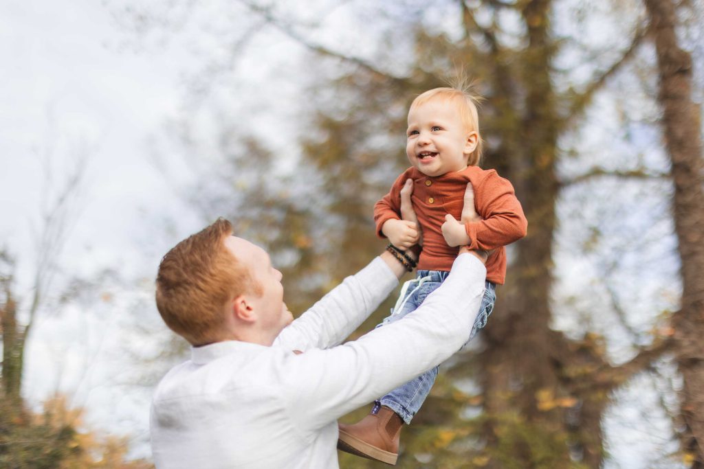 During a delightful family mini-session at Belair Mansion, an adult in a white shirt lifts a smiling toddler in an orange shirt and blue jeans against the backdrop of lush trees.
