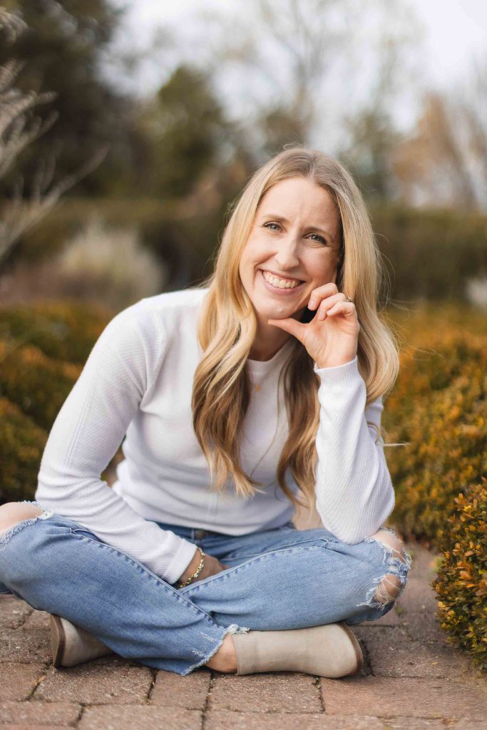 A woman with long blonde hair wearing a white sweater and ripped jeans sits cross-legged on a brick path outdoors, smiling and resting her chin on her hand. Surrounded by shrubs and trees, she exudes warmth as if waiting for family at the picturesque Belair Mansion.